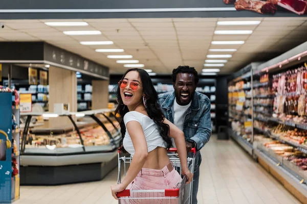 Glückliche asiatische Mädchen mit Sonnenbrille, die in die Kamera schaut, während sie im Einkaufswagen in der Nähe positiver afrikanisch-amerikanischer Mann sitzt — Stockfoto