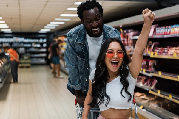 Happy asian girl in sunglasses gesturing while sitting in shopping cart near positive african american man — Stock Photo