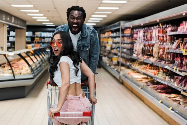 Surprised asian woman in sunglasses sitting in shopping cart near positive african american man — Stock Photo