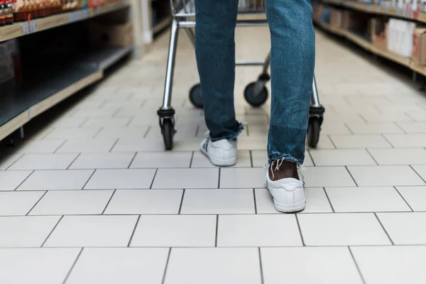 Cropped view of african american man in blue denim jeans walking with shopping cart — Stock Photo