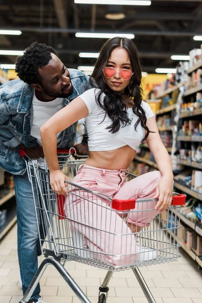 Handsome african american man looking at stylish asian girl in sunglasses sitting in shopping cart in store — Stock Photo