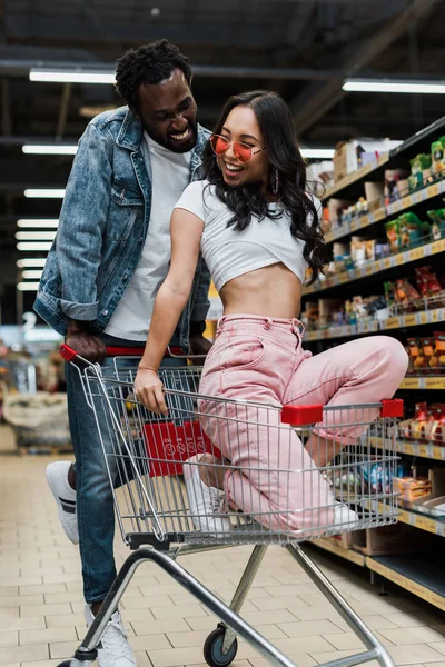 Красивий афроамериканський чоловік дивиться на стильну азіатську жінку в сонцезахисних окулярах, сидячи в кошику в магазині — стокове фото