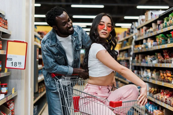 Handsome african american man looking at stylish asian woman in sunglasses sitting in shopping cart — Stock Photo