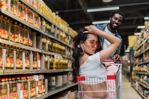 Enfoque selectivo de la elegante chica asiática en gafas de sol cerca de hombre afroamericano feliz en la tienda - foto de stock