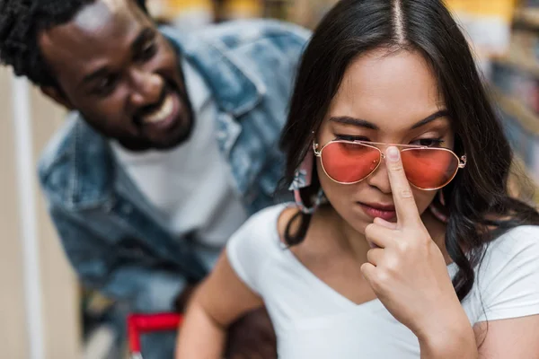 Foco seletivo de mulher asiática tocando óculos de sol perto de homem americano africano feliz — Fotografia de Stock