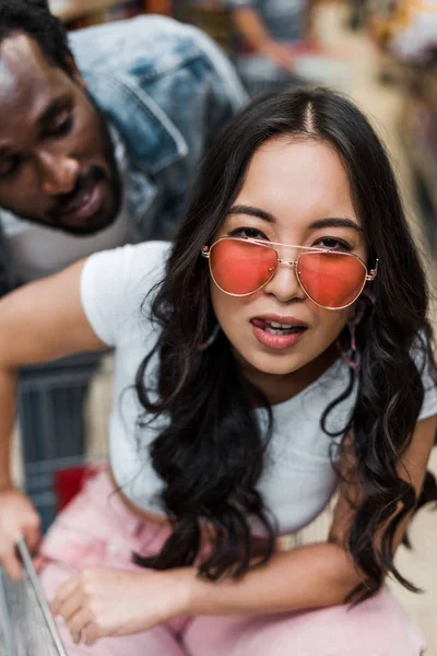 Mise au point sélective de fille asiatique dans des lunettes de soleil en regardant la caméra près de l'homme afro-américain — Photo de stock