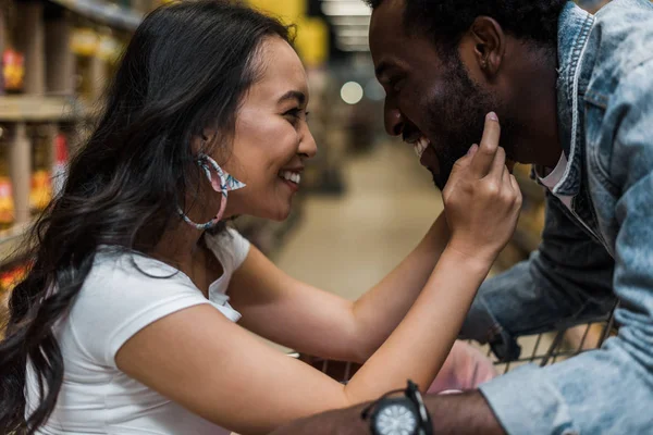 Selektiver Fokus glücklicher asiatischer Mädchen, die Gesicht eines gutaussehenden und fröhlichen afrikanisch-amerikanischen Mannes berühren — Stockfoto