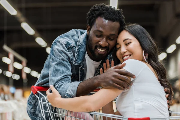 Heureux afro-américain homme étreignant asiatique jolie femme avec les yeux fermés en magasin — Photo de stock