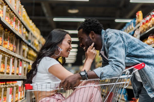 Happy asian girl sitting in shopping cart and touching face of handsome and cheerful african american man — Stock Photo