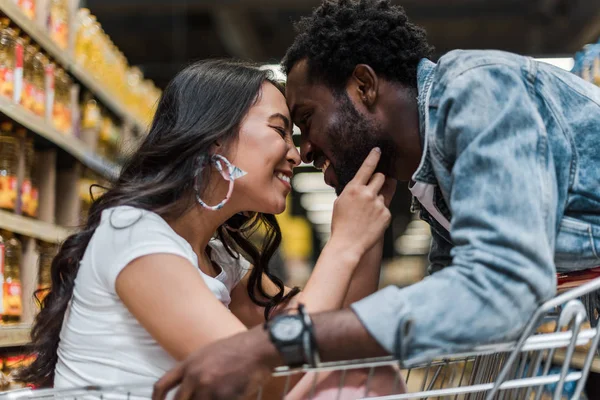 Selective focus of cheerful asian girl sitting in shopping cart and touching face of handsome and happy african american man — Stock Photo