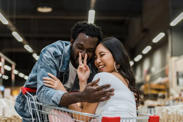 Foco seletivo de alegre asiático menina sentado no carrinho de compras e tocando nariz africano americano homem — Fotografia de Stock