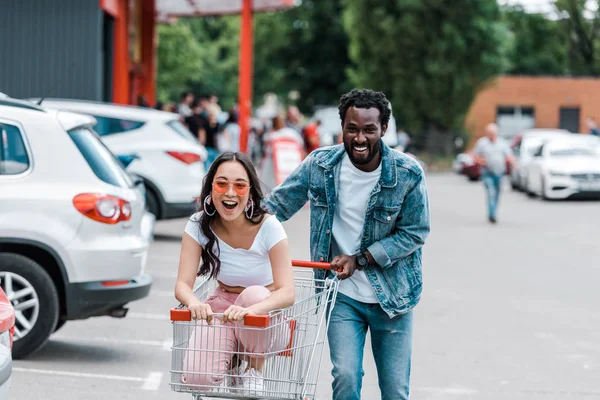 Happy african american man walking while asian girl in sunglasses sitting in shopping cart — Stock Photo