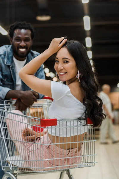 Foco seletivo de alegre asiático menina sentado no carrinho de compras perto bonito africano americano homem — Fotografia de Stock