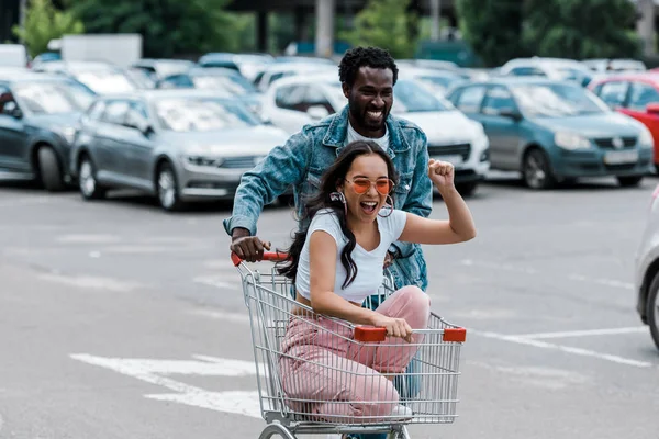 Щаслива азіатська дівчина жестикулює, сидячи у візку біля афроамериканського чоловіка та автомобілів — стокове фото