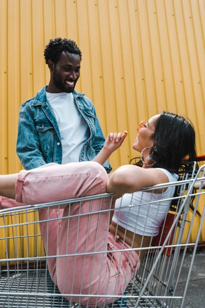 Foco seletivo da menina em óculos de sol sentado no carrinho de compras perto de homem americano africano fora — Fotografia de Stock