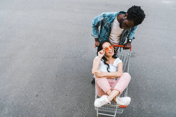 Opinião aérea o homem do americano africano que olha a menina asiática feliz que senta-se no trole da compra e nos óculos de sol de toque — Fotografia de Stock