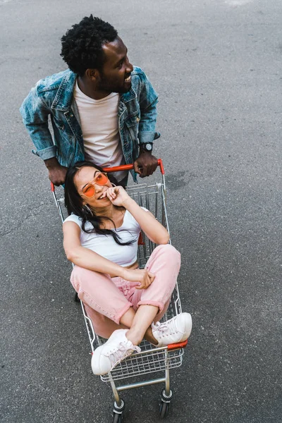 Vista aérea do homem do americano africano que está perto da menina asiática feliz que senta-se no trole da compra fora — Fotografia de Stock