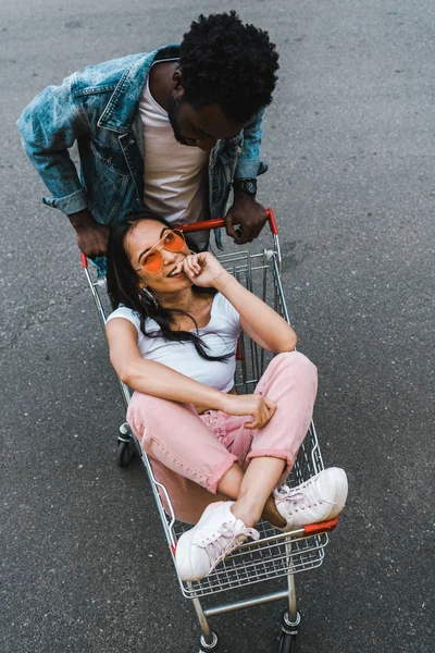 Вид зверху на афроамериканця, який дивиться на щасливу азіатську дівчину, що сидить на шопінгу зовні — стокове фото