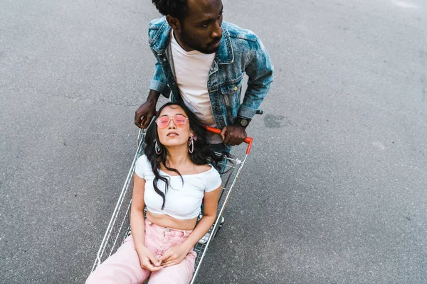 Vue aérienne de l'homme afro-américain debout près de la fille asiatique assis dans le chariot de magasinage à l'extérieur — Photo de stock