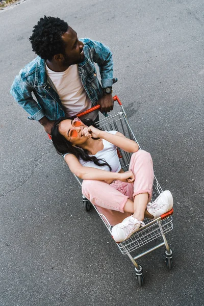 Vista aerea di afro americano uomo in piedi vicino asiatico ragazza seduta in shopping trolley e copertura faccia al di fuori — Foto stock