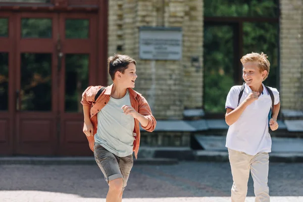 Dos colegiales lindos corriendo en el patio de la escuela mientras sonríen y se miran - foto de stock