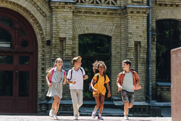 Cuatro escolares multiculturales alegres sonriendo mientras corren en el patio de la escuela - foto de stock