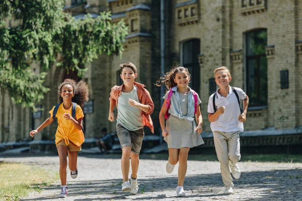 Four happy multiethnic schoolchildren smiling while running in schoolyard — Stock Photo
