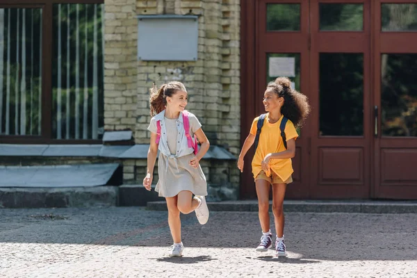 Дві веселі багатоетнічні школярки посміхаються під час бігу на шкільному подвір'ї — стокове фото