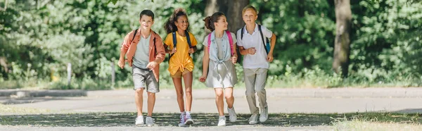 Panoramaaufnahme von vier fröhlichen multikulturellen Schulkindern beim Laufen im sonnigen Park — Stockfoto