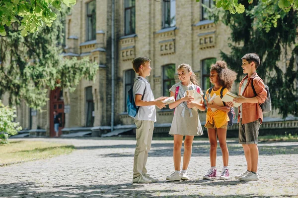 Quatro estudantes multiculturais bonitos falando enquanto estão em pé no pátio da escola e segurando livros — Fotografia de Stock