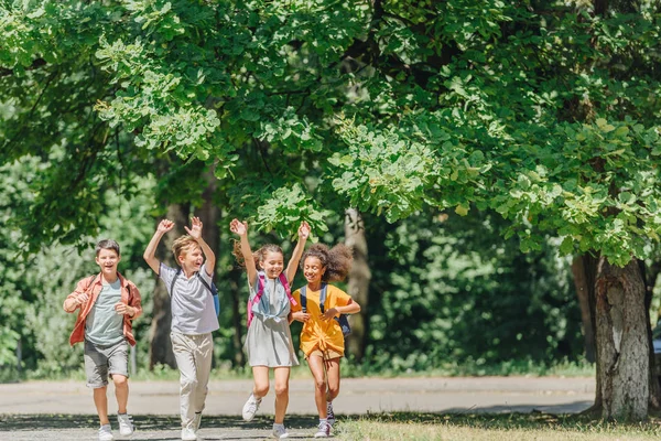 Four happy multicultural schoolkids gesturing with raised hands while running in park — Stock Photo