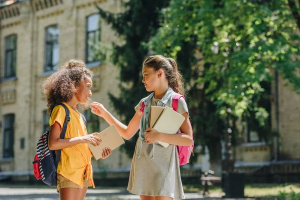 Two multicultural schoolgirls holding books and speaking while standing in schoolyard — Stock Photo
