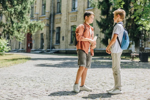 Two cute schoolboys holding books and speaking while standing in schoolyard — Stock Photo