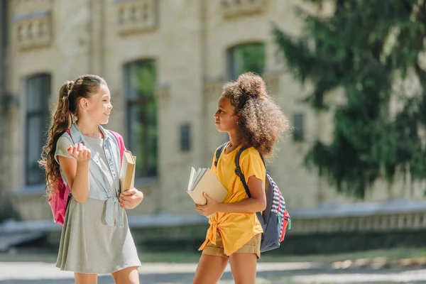 Две весёлые мультикультурные школьницы, держащие книги и разговаривающие во дворе школы — стоковое фото
