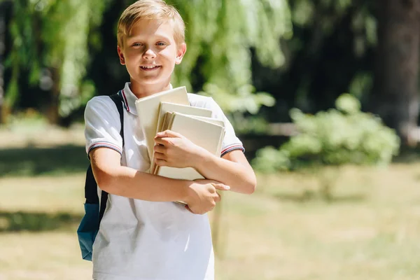 Entzückender Schuljunge mit Rucksack, der Bücher hält und in die Kamera lächelt — Stockfoto