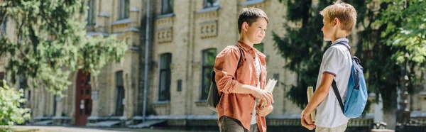 Panoramic shot of two cute schoolboys speaking while holding books in schoolyard — Stock Photo