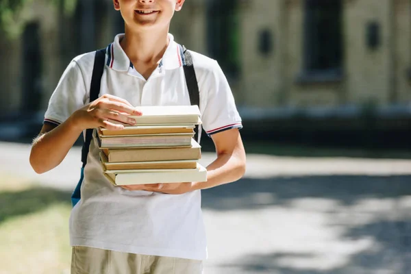 Обрезанный вид улыбающегося школьника с рюкзаком, держащим книги во дворе школы — стоковое фото
