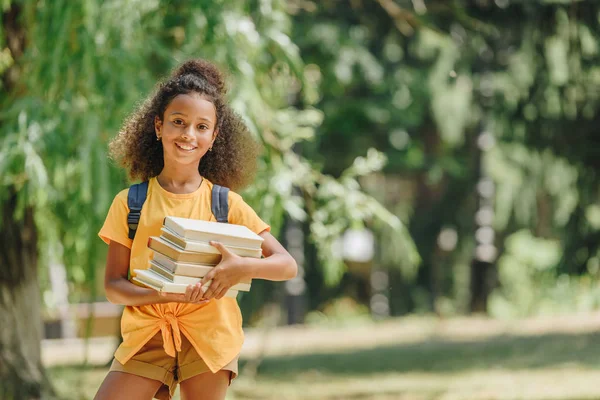 Joyeuse écolière afro-américaine tenant des livres et souriant à la caméra dans le parc — Photo de stock