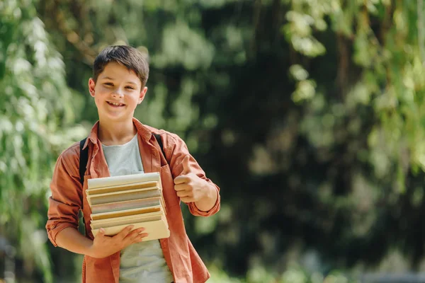 Menino de escola alegre sorrindo para a câmera enquanto mostra o polegar para cima e segurando livros — Fotografia de Stock