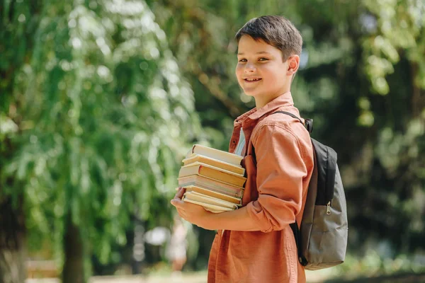 Feliz colegial sonriendo a la cámara mientras está de pie en el parque y sosteniendo libros - foto de stock