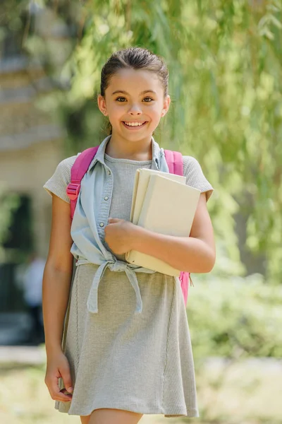 Niedliches, lächelndes Schulmädchen, das in die Kamera schaut, während es Bücher im Park hält — Stockfoto