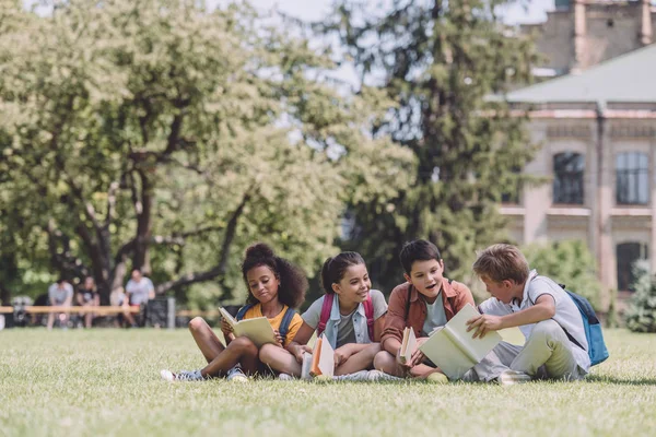 Quatre écoliers multiculturels joyeux assis sur la pelouse et lisant des livres — Photo de stock