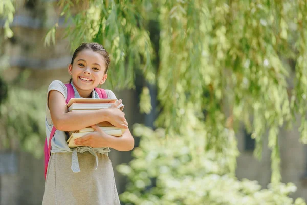 Adorable, happy schoolgirl looking at camera while holding books in park — Stock Photo