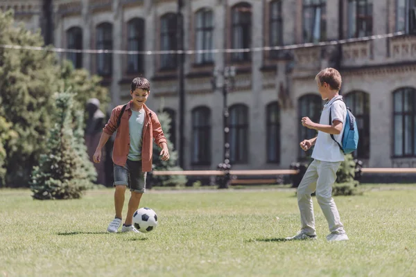 Deux écoliers joyeux avec des sacs à dos jouant au football sur la pelouse près de l'école — Photo de stock