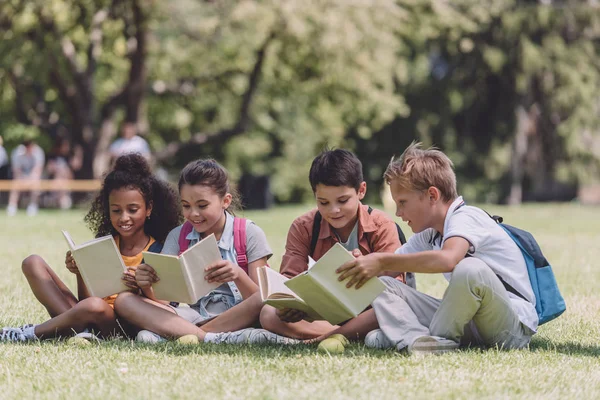 Cuatro adorables escolares multiculturales sentados en el césped y leyendo libros - foto de stock