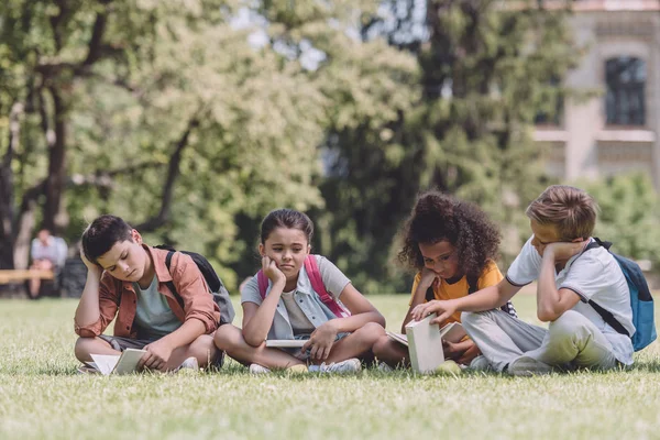 Quatro escolares multiculturais entediados com livros sentados no gramado no parque — Fotografia de Stock