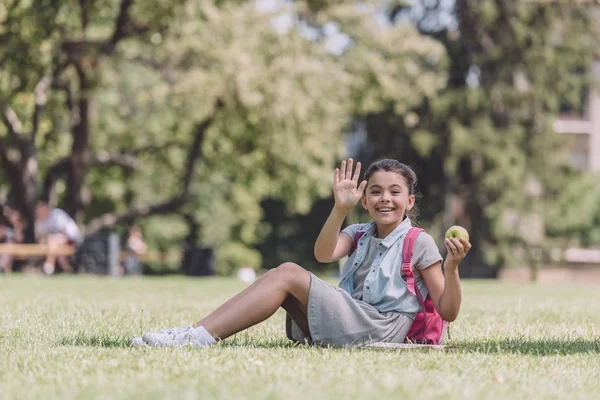 Cheerful schoolgirl waving hand while sitting on lawn in park and looking at camera — Stock Photo