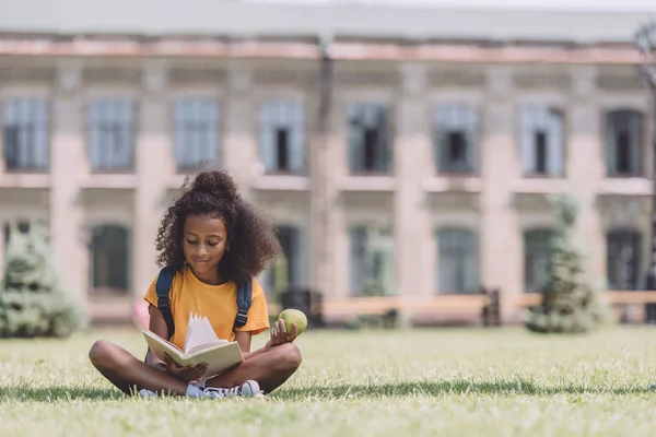 Улыбающаяся африканская американская школьница держит яблоко и читает книгу, сидя на лужайке — стоковое фото