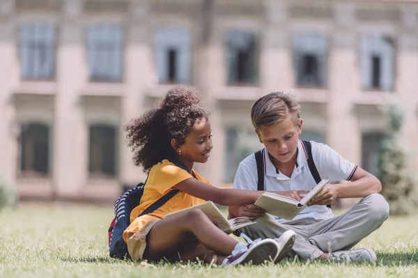 Dos adorables escolares multiculturales sentados en el césped y leyendo libros juntos - foto de stock