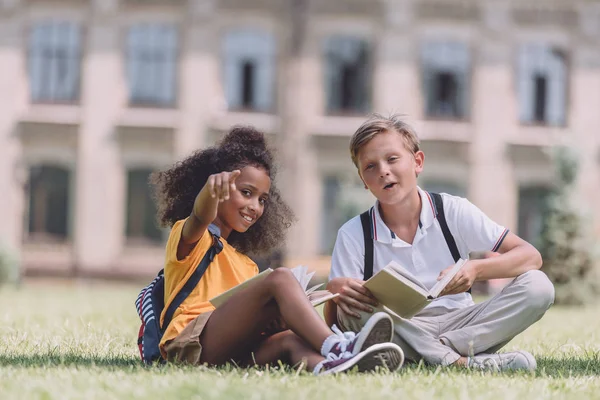 Веселий афроамериканець школярка дивиться на камеру і вказуючи пальцем, сидячи на галявині біля багатонаціонального друга — стокове фото